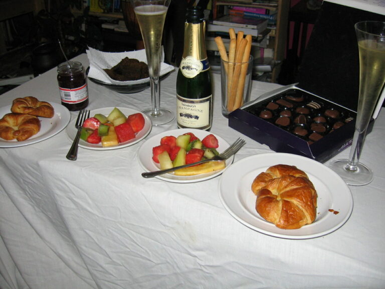 Auf einem Tisch mit weißer Tischdecke sind Obstteller, eine Pralinenschachtel und eine Champagnerflasche neben gefüllten Gläsern zu sehen